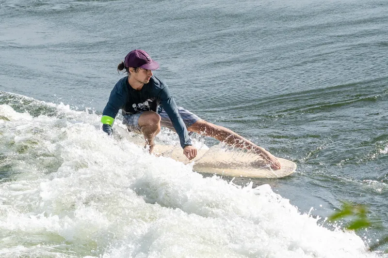 Un surfeur en casquette mauve, accroupi sur sa planche, frôle l'écume de la main