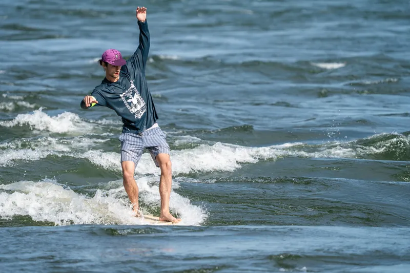 Un surfeur en casquette mauve, un bras levé, debout sur la vague