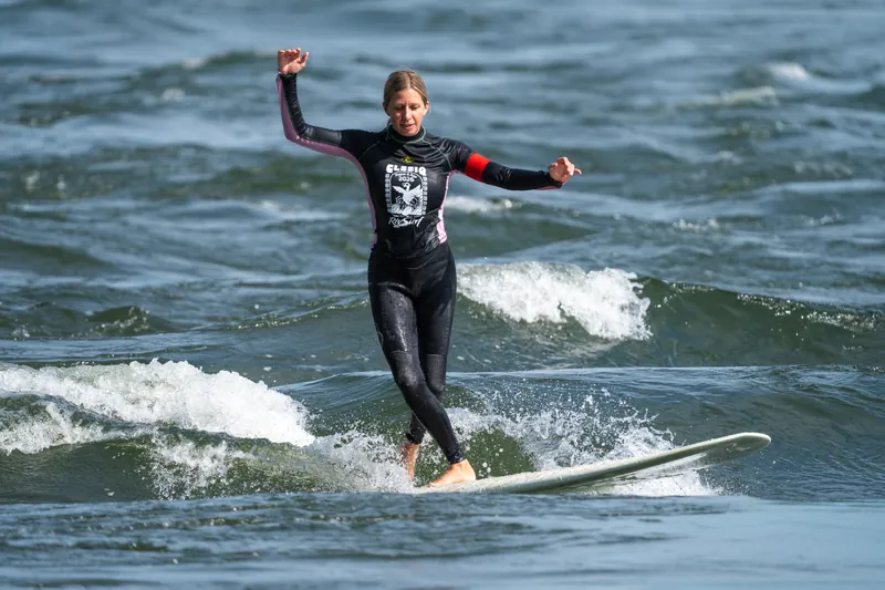 Une surfeuse en combinaison noire, bras ouverts, marche sur son longboard
